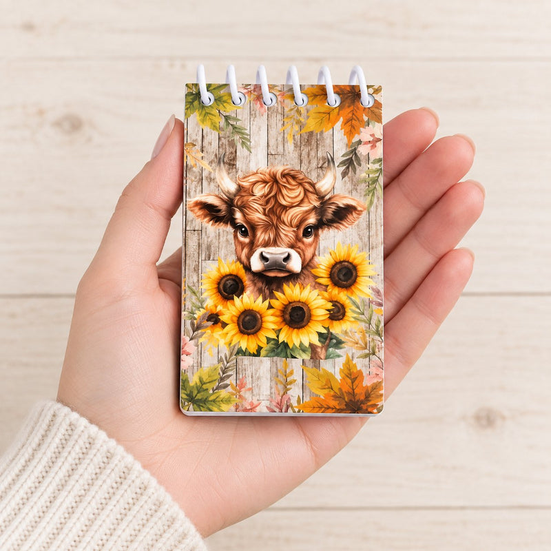 Hand holding a pocket-sized Highland cow spiral notebook with a sunflower cover