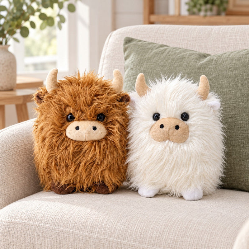 Brown and white shaggy Highland cow plush toys together on an ivory sofa, showing both 22 cm colour options. Each plush sold separately.