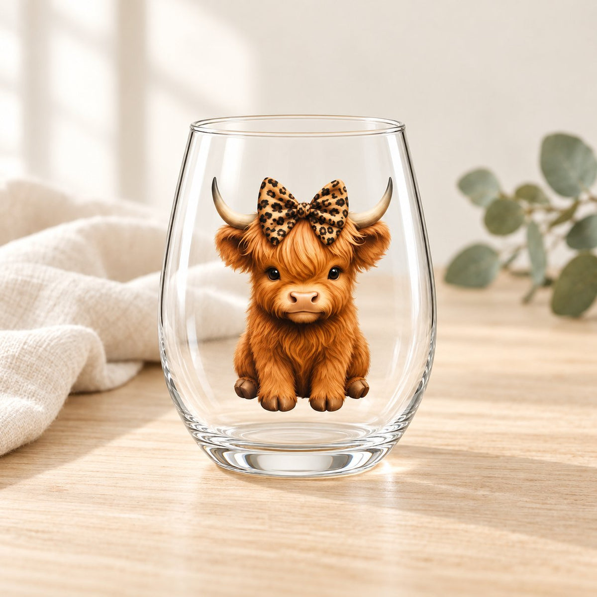 Leopard-bow Highland cow glass styled on pale timber with ivory linen and eucalyptus; props are not included.