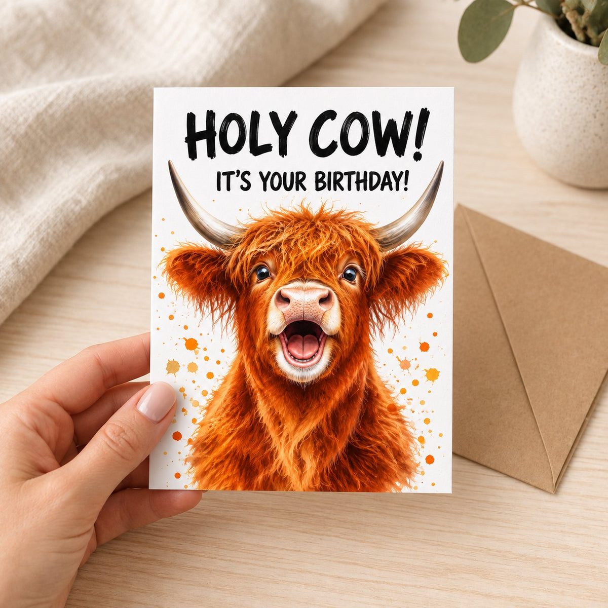Hand holding the Holy Cow birthday card with its kraft envelope nearby; linen and plant are styling props.