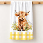 Meadow Calf Highland cow tea towel with meadow flowers and a yellow gingham border, shown folded on a pale timber rail