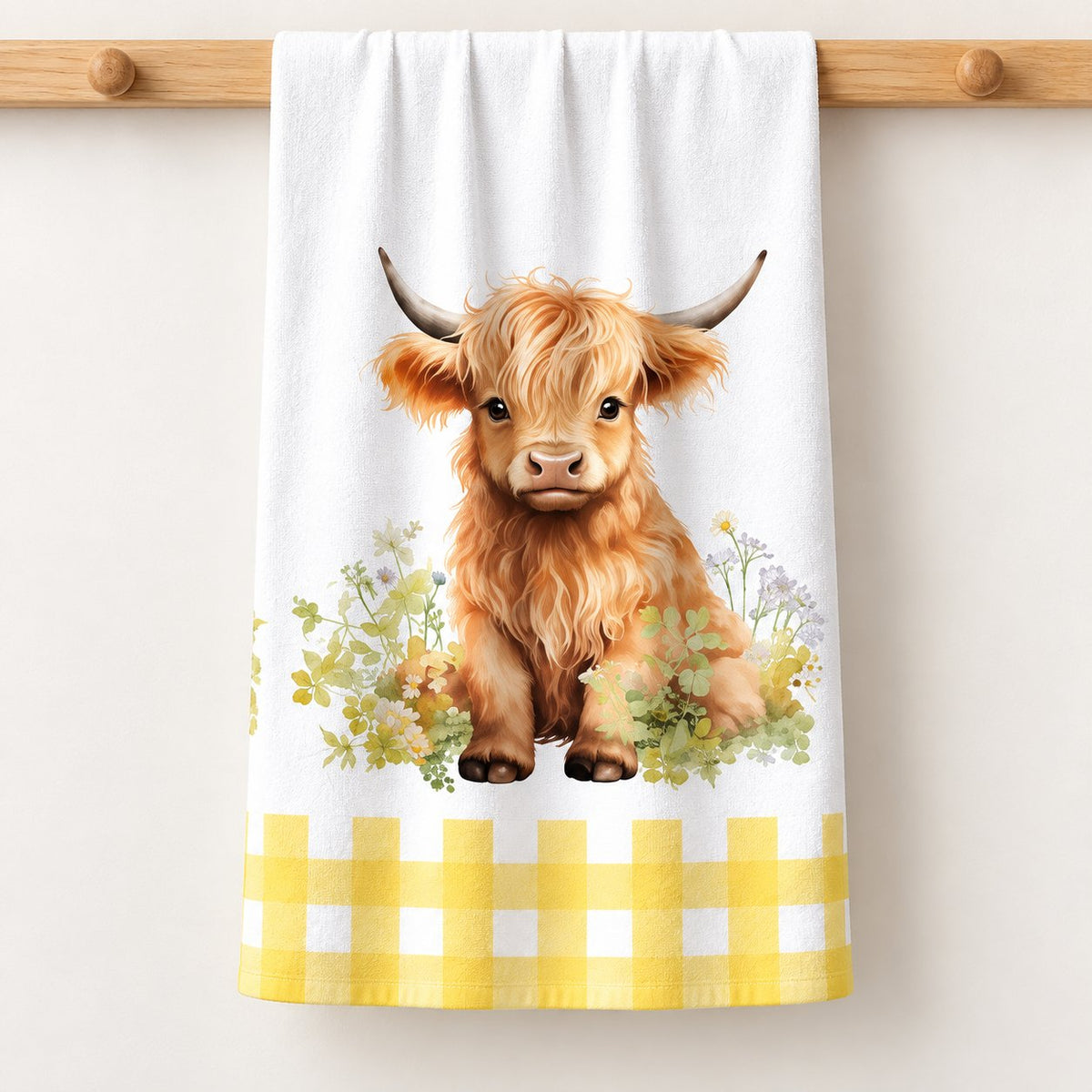 Meadow Calf Highland cow tea towel with meadow flowers and a yellow gingham border, shown folded on a pale timber rail