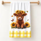 Sunflower Calf Highland cow tea towel with butterflies and a yellow gingham border, shown folded on a pale timber rail